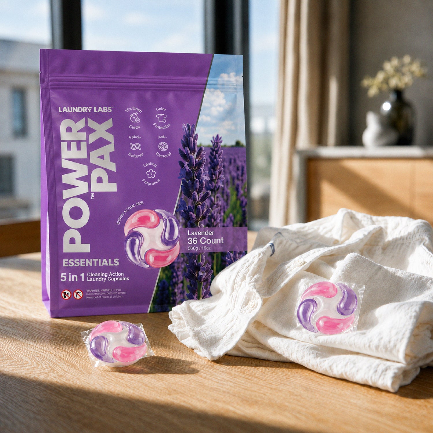 A stand-up pouch of Laundry Labs Power Pax Essentials laundry detergent pods stands on a wooden table beside a white towel and a single loose pod. The package is predominantly purple with lavender imagery and product text, and the scene is lit by warm natural light from a nearby window.